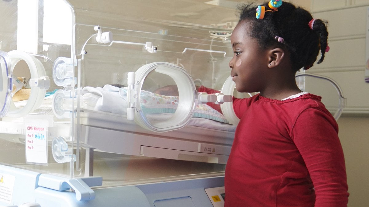 A young girl standing by a baby incubator in a hospital environment