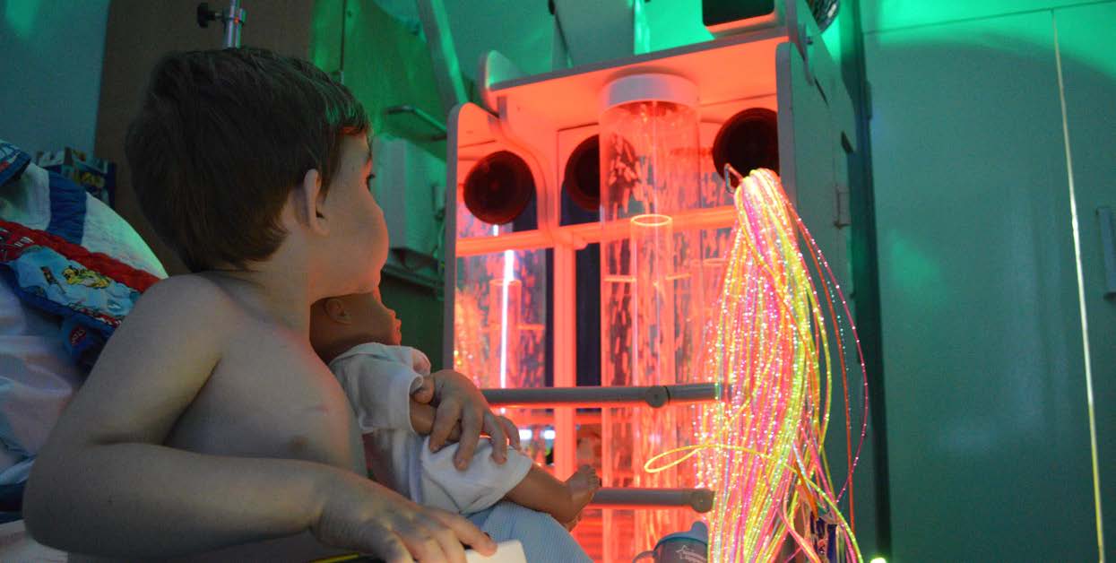 A young boy in a hospital bed with a toy baby, sitting in front of sensory equipment